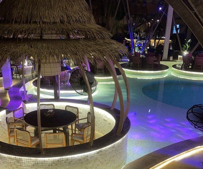 An evening view of a resort or restaurant featuring a swimming pool with illuminated edges, surrounded by seating areas with thatched roofs and decorative lighting.