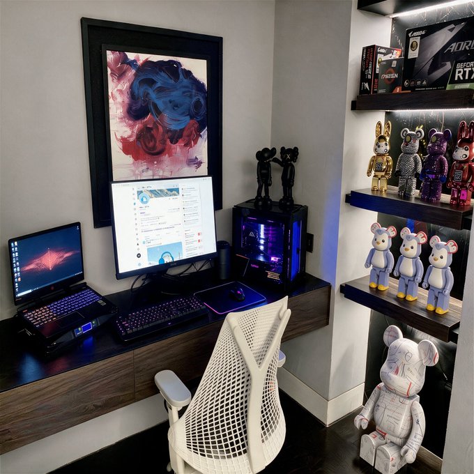 A photograph of a modern gaming/tech setup featuring a curved monitor, laptop, RGB keyboard, gaming PC, a white ergonomic chair, Bearbrick figurines on shelves, and framed abstract art.