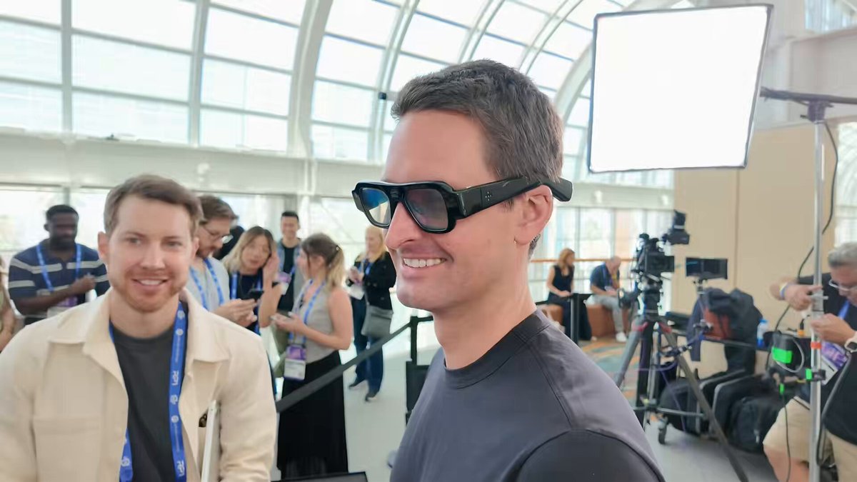 In the image, there are two men standing next to each other with one wearing black sunglasses and a gray shirt while the other has a white jacket on. They appear to be posing for a photo or participating in an event. The background shows several people gathered around them, some of whom have cameras set up to capture their presence. A sign is also visible in the scene that reads "The eyewear equivalent of implementing stablecoins into your Bitcoin app." This phrase seems unrelated to the main subjects and could be a reference or joke related to the event they are attending.