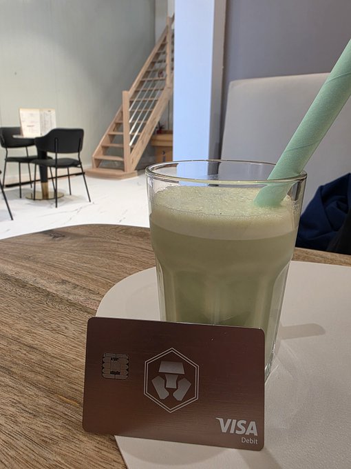 A glass of green matcha drink with a green straw on a table next to a dark VISA Debit card with a Crypto.com logo, with a staircase and chairs in the background.