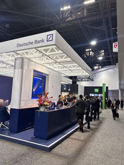 A view of a Deutsche Bank booth at a financial conference, featuring a large white sign with the company's blue logo above a staffed reception desk decorated with flowers.