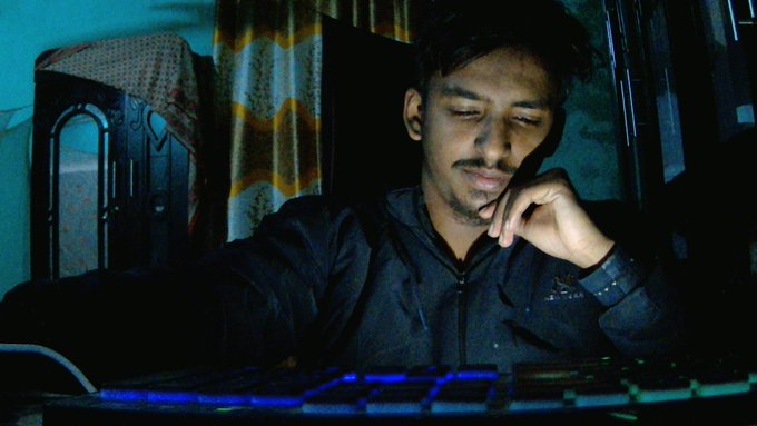 The image shows a man sitting at a computer desk, wearing a black jacket or hoodie while using a keyboard. The background features a window that allows natural light into the room. The photo has been edited with a filter applied, giving it a blue tint. The caption reads "Good evening x fam i am back What makes me most optimistic about @useTria is where they could be in the next year or two. By combining true self-custody with a smooth and practical user experience, Tria is".