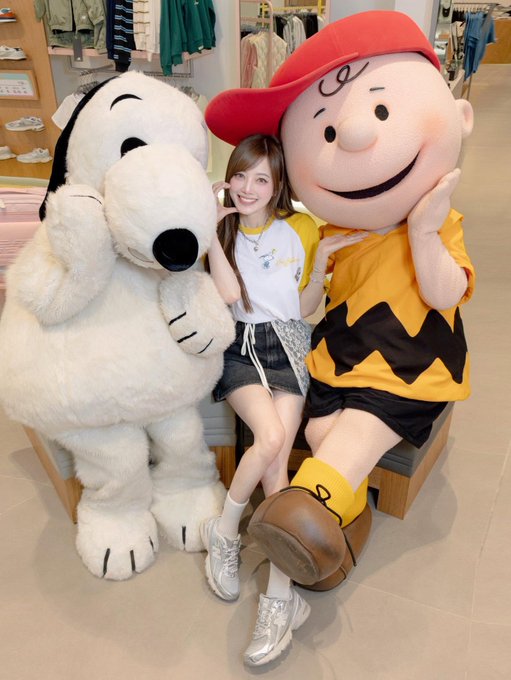 A woman smiles while posing between a large Snoopy mascot and a Charlie Brown mascot in what appears to be a clothing store.