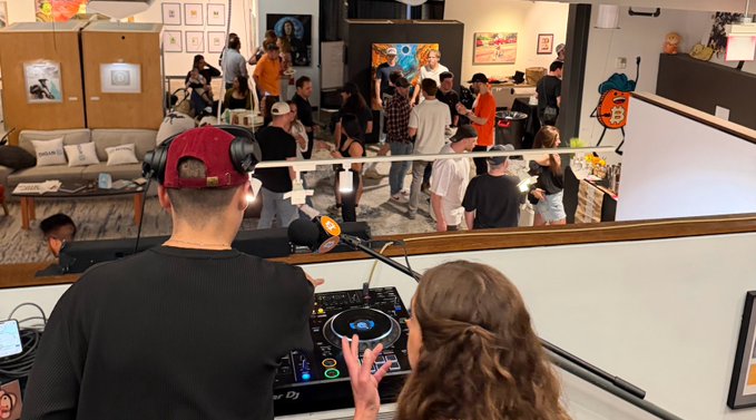 The image shows a man wearing a red baseball cap, standing behind a DJ booth with his hands up, possibly playing music or adjusting equipment. A woman stands next to him, looking at something above them. The scene takes place inside an art gallery where several people are gathered around and observing the artwork on display. In the background, there is a painting hanging on the wall that adds to the artistic atmosphere of the space.