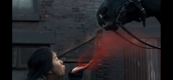 A woman with dark hair exhales a stream of red mist or smoke from her mouth, reaching her hand toward a dark horse whose head is visible above her, set against a brick wall background.