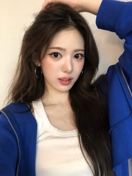 A close-up portrait of a young woman with long dark hair, wearing a blue zip-up jacket over a white top, posing with one arm raised above her head.