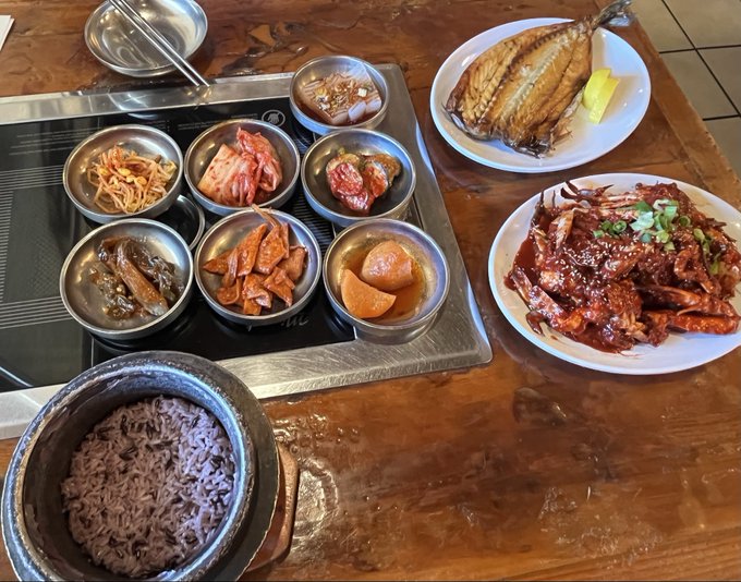 A Korean meal spread featuring grilled mackerel, spicy braised crab, purple rice in a stone bowl, and various banchan side dishes in metal bowls on a wooden table.