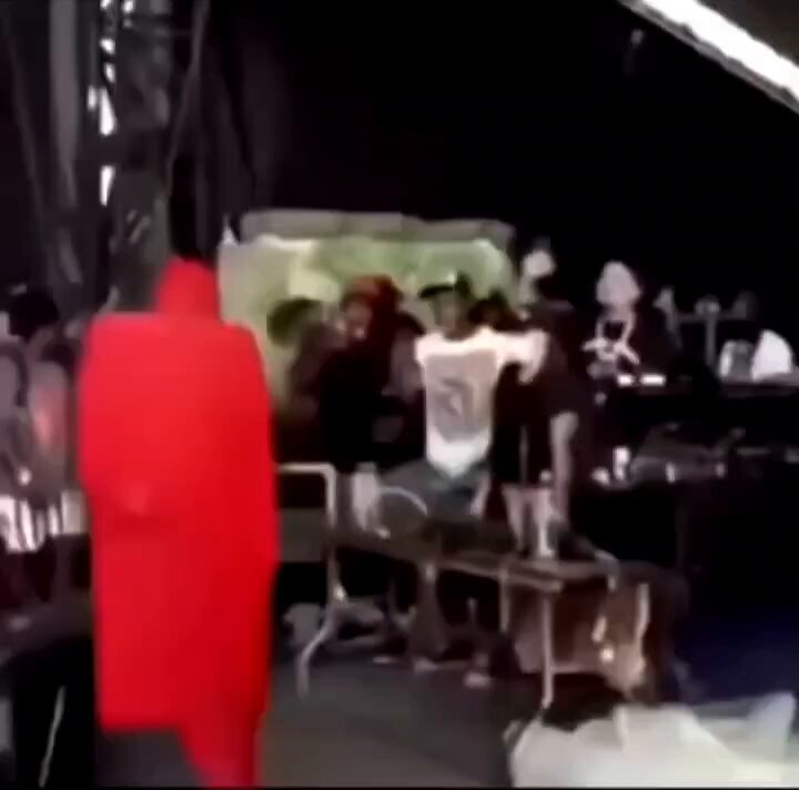 The image shows a group of people gathered around an event, with some standing near a stage while others are sitting or standing behind a bench. A person wearing a red outfit stands out among them. In the background, there's a sign that reads "POV: You finally buy the token The next candle:". This suggests that the image is taken from a perspective where the viewer can see the event unfold and understand its context better.