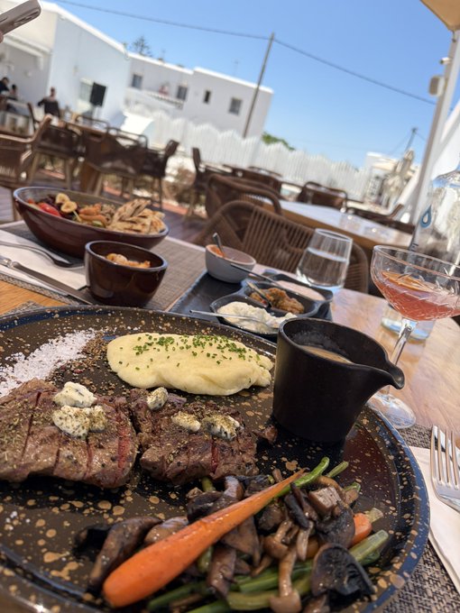A gourmet meal featuring steak, mashed potatoes, and vegetables is served at an outdoor restaurant in Mykonos, paired with a pink cocktail. The Mediterranean setting with whitewashed buildings in the background complements the caption "Mykonos Maxxxing," highlighting a luxurious dining experience on the island.