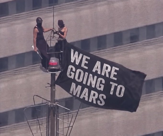 Two masked protesters unfurl a large black banner reading "WE ARE GOING TO MARS" from atop a flagpole or street fixture, embodying the post's "Bullish on Mars" sentiment with a literal proclamation of optimism about humanity's future on the Red Planet.