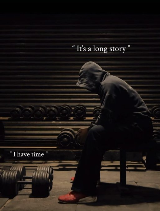 The image features a man sitting on a bench, wearing a hoodie and red shoes while holding two dumbbells. The background includes a wall with a shutter door and several dumbbells arranged behind him. There are also text elements in the image that read "It's a long story" and "I have time".