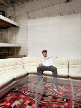 A young man wearing a 'CELINE PARIS' sweatshirt and black beanie sits on a plush white sectional sofa in a loft-style space with a glass coffee table and patterned rug.