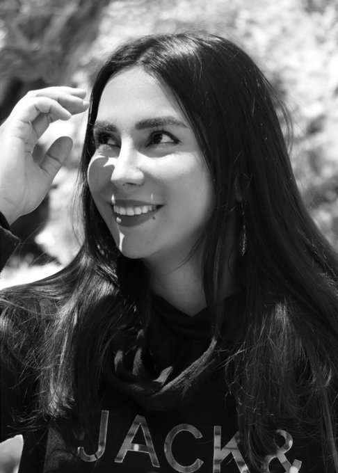 A black-and-white portrait of a smiling young woman with long dark hair, wearing a 'JACK &' hoodie, with her hand raised near her head in a candid outdoor shot.