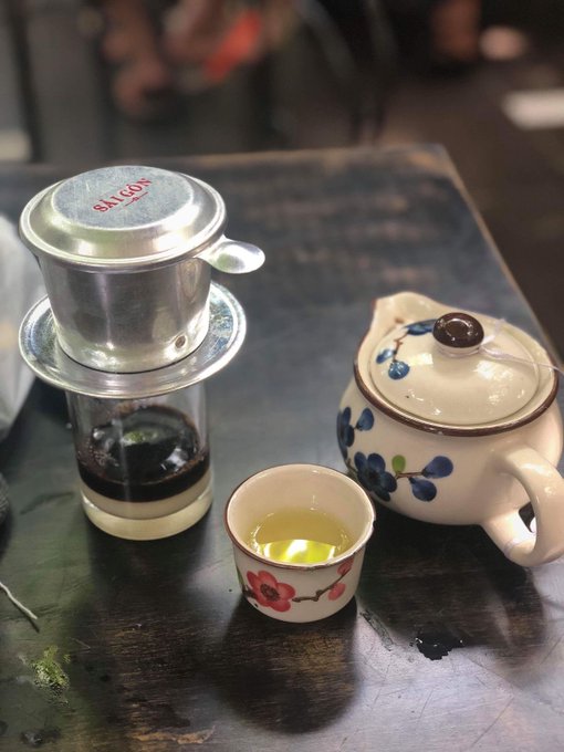 A traditional Vietnamese coffee setup on a dark wooden table, featuring a silver Saigon phin drip filter over a glass, a small floral teacup with brewed tea, and a blue and white floral teapot.