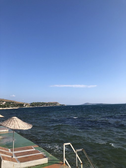 A serene seaside view shows a wooden deck with a thatched umbrella and metal railing overlooking a deep blue sea, with a distant coastline and clear sky in the background. The image of a calm horizon contrasts with the turbulent crypto market losses mentioned in the post about Bitcoin at $72k and altcoin holdings.