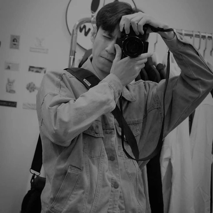 A black and white photo of a man wearing a jacket, holding a camera up to his face as if taking a picture, with clothing visible on a rack behind him.