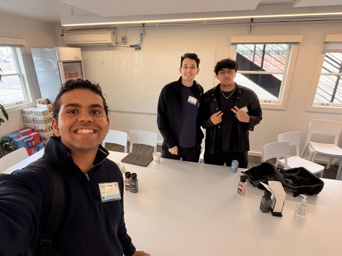 Three young men pose for a selfie-style picture around a white table in a bright meeting room or classroom setting.