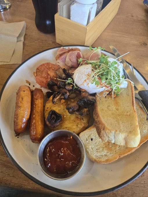 A hearty breakfast plate on a wooden table featuring two sausages, bacon, roasted tomatoes, mushrooms, poached eggs garnished with microgreens, toasted bread, and a small bowl of ketchup.