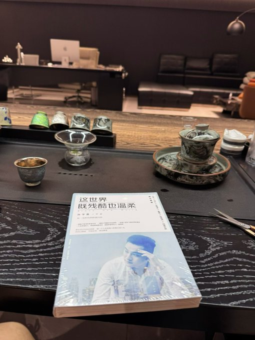 A book titled "这世界既残酷也温柔" by 刘擎 rests on a wooden table set with a traditional tea ceremony, with a modern office visible in the background.