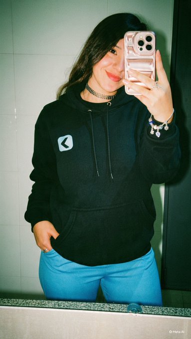 A person takes a mirror selfie wearing a black hoodie with a logo, blue pants, and a choker necklace while holding a pink phone case.