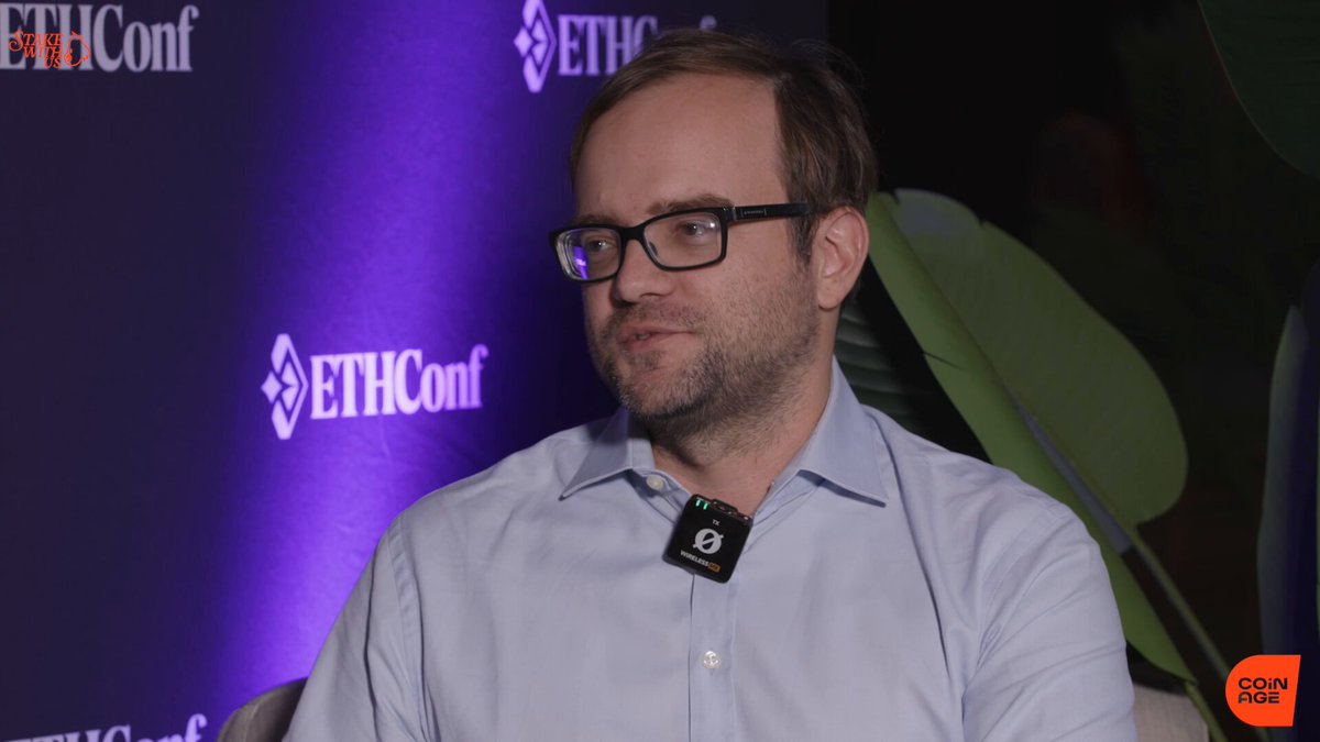 The image features a man wearing glasses, sitting down with his hands resting on his lap. He appears to be engaged in conversation or listening attentively as he looks off into the distance. The background of the image includes a purple wall adorned with logos for "ETHCONF" and "Lighter_xyz".