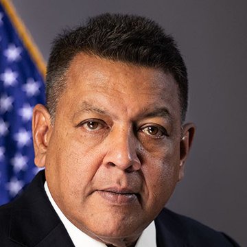 A formal portrait shows Mel Gunewardena, a U.S. CFTC official, in professional attire with an American flag visible in the background, aligning with his role in advocating for U.S. leadership in tokenization, stablecoins, smart contracts, and AI as described in the accompanying post.