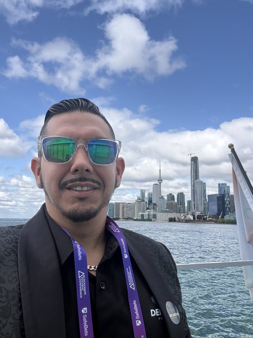 A man wearing green-reflective sunglasses and a black blazer poses for a selfie with the Toronto skyline and CN Tower visible across the water behind him.