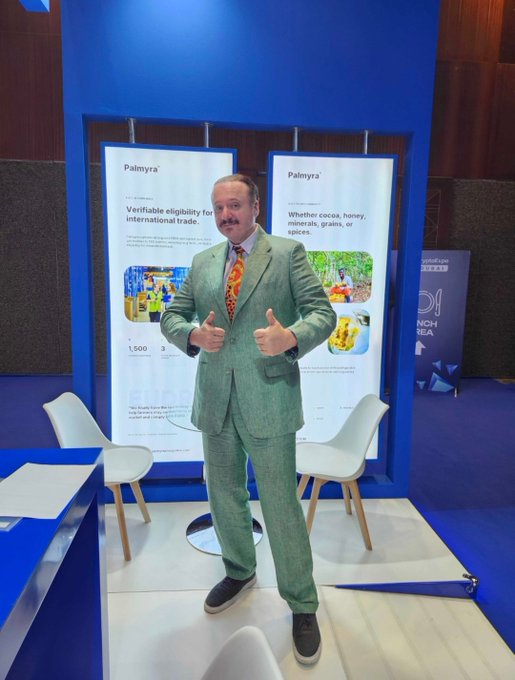 A man with a mustache wearing a green suit and orange patterned tie giving two thumbs up at what appears to be a Palmyra trade exhibition booth.