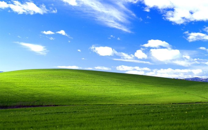 A vibrant green rolling hill stretches beneath a bright blue sky dotted with fluffy white clouds, evoking the simple joy of "touching the grass." The iconic landscape, reminiscent of the classic Windows XP "Bliss" wallpaper, captures a peaceful, idyllic moment in nature.