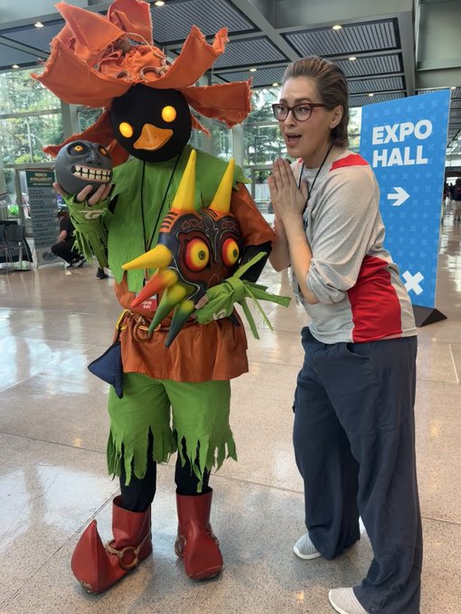 A convention hall scene featuring a cosplayer dressed as Skull Kid from The Legend of Zelda wearing a Majora's Mask, posing alongside a woman in a gray shirt near an 'EXPO HALL' directional sign.