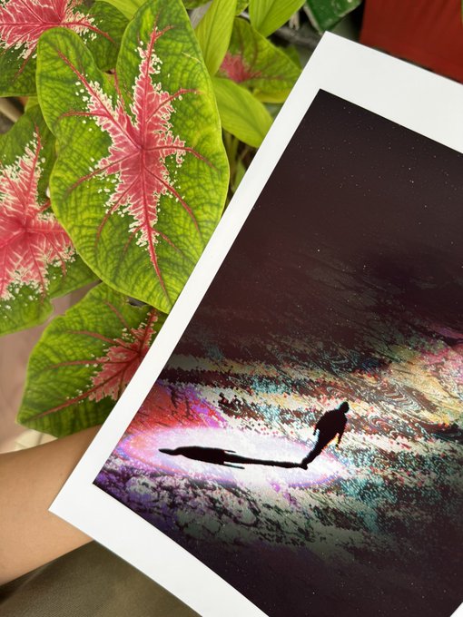 A photograph shows a person holding a printed artwork depicting silhouetted figures walking across a vibrant, cosmic landscape, with pink and green caladium plants visible in the background.