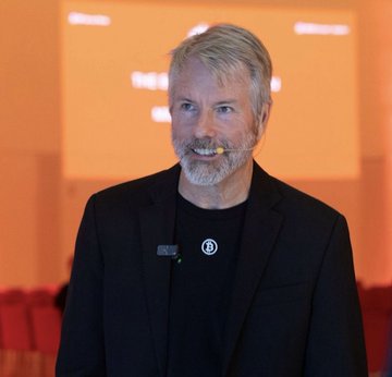 A portrait of a smiling man with gray hair and a beard, wearing a black shirt with a circular pin, photographed against a warm-toned indoor background.