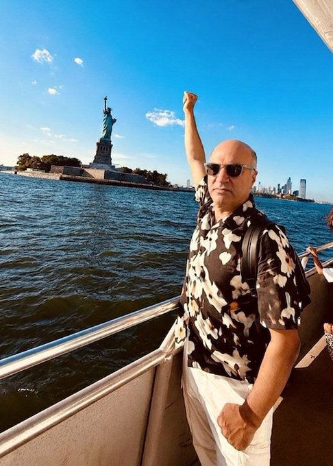 A bald man wearing sunglasses and a black and white patterned shirt, posing on a boat with his fist raised with the Statue of Liberty in the background.