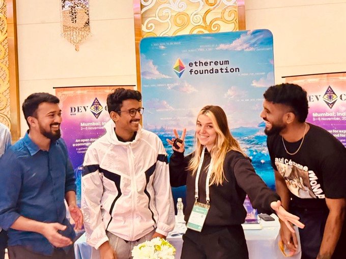 Four young people pose cheerfully in front of an Ethereum Foundation banner at a Devcon event in Mumbai, with one person flashing a peace sign, capturing the excitement of joining the Devcon India Ecosystem team for Ethereum's flagship conference returning to India after over a decade.