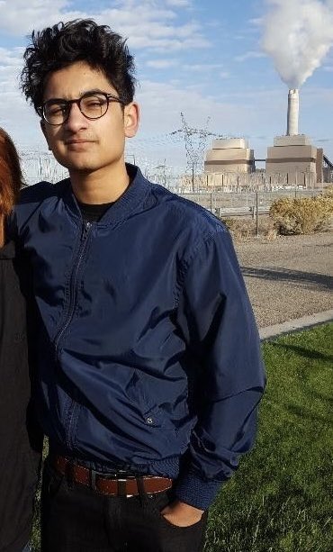 A young man wearing glasses and a blue bomber jacket poses for a photo outdoors, with a power plant releasing steam visible in the background.