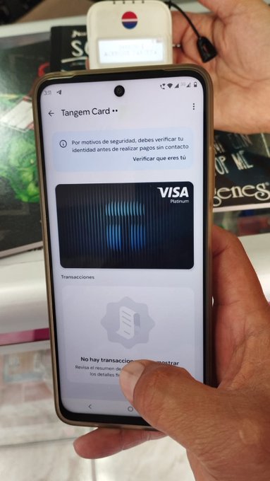 A person holds a smartphone displaying the Tangem Card app in Spanish, showing a Visa Premium virtual card and a verification prompt; a Tangem hardware wallet is visible behind the phone.