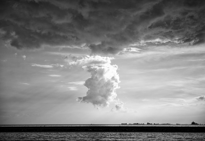 A black and white photograph of a dramatic cumulus cloud formation rising above a body of water, with a distant shoreline visible on the horizon.