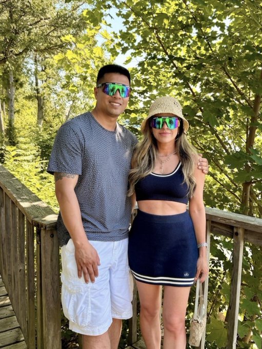 A couple posing together outdoors; the man wears a grey shirt and white shorts, while the woman wears a navy blue two-piece outfit and a straw hat, both sporting matching colorful sunglasses.