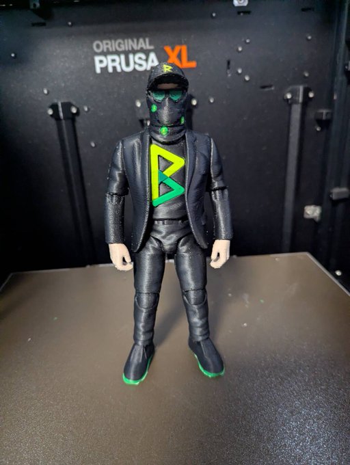 A 3D-printed action figure stands on a surface in front of an 'ORIGINAL PRUSA XL' 3D printer; the figure wears a black suit with a green 'R' logo and matching accessories.
