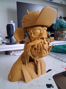 A clay sculpture of a Bored Ape-style character wearing a pirate hat and an eyepatch, displayed on a desk with a monitor in the background.