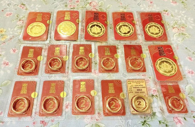 Several packaged gold coins and bars, including 'MINH TAM BAC' and '999.9 FINE GOLD' items, are arranged on a floral patterned surface.