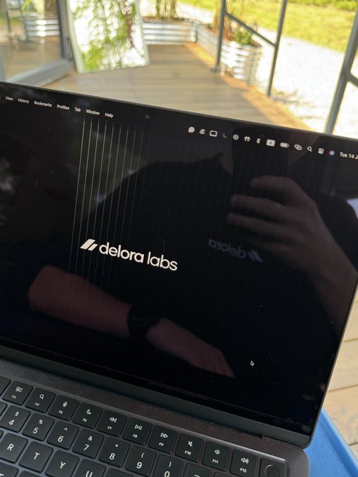 A laptop screen displays the logo for "delora labs" on a dark background, with a reflection of the photographer visible on the screen, set against an outdoor patio area.