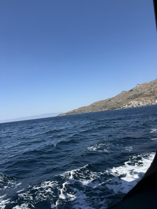 A boat cuts through deep blue sea waters, leaving white wake behind, with a coastal town visible on a distant hillside under a clear sky. The image contrasts the post's discussion of Fhenix's blockchain privacy infrastructure by offering a serene, analog moment of travel across open water, perhaps symbolizing exploration or uncharted territory.