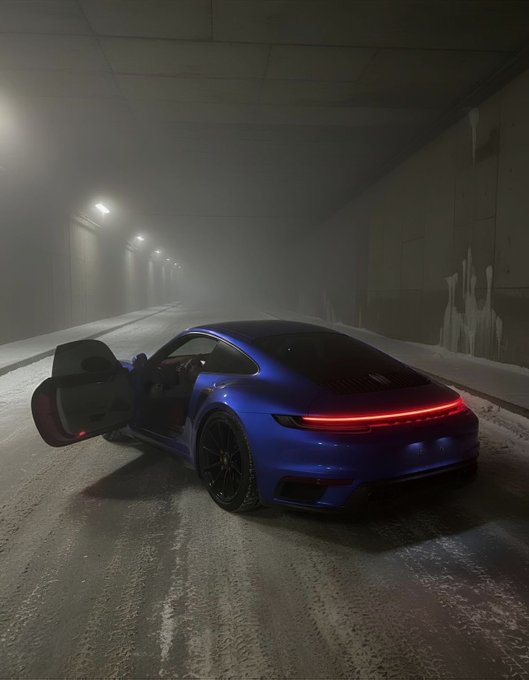 A blue Porsche sports car with its door open, parked in a foggy tunnel with the rear LED light bar illuminated, creating a moody cinematic atmosphere.