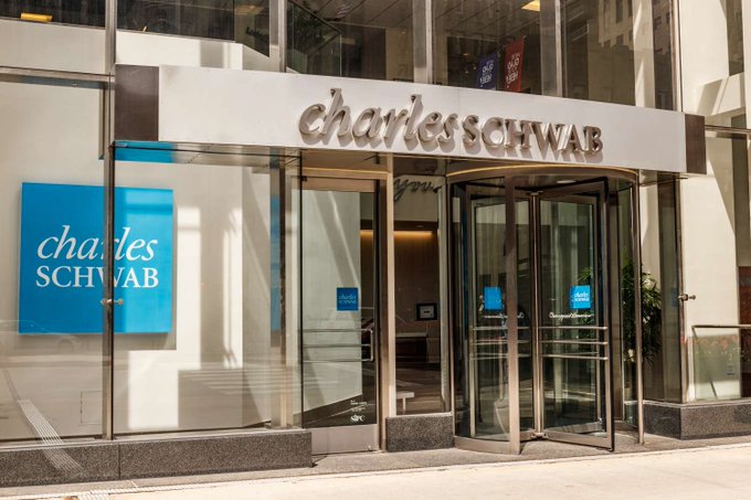 The image shows the entrance to a Charles Schwab branch, featuring the company's name in large lettering above a revolving glass door and a blue "charles schwab" sign in the window. It depicts the brokerage firm referenced in the breaking news that Charles Schwab, with $13.1 trillion in client assets, will begin offering Solana ($SOL) trading to its clients.