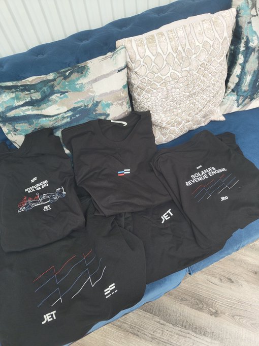 Black JET-branded t-shirts, hoodies, and long-sleeve shirts are laid out on a blue tufted couch with decorative pillows, featuring logos and text like 'SOLANAS REVENUE ENGINE' and 'ACCELERATING DEX APES'.