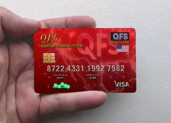 A hand holding a red 'QFS Quantum Financial System' Visa debit card with the number 8722 4331 1992 7982 and a US flag printed on the card.