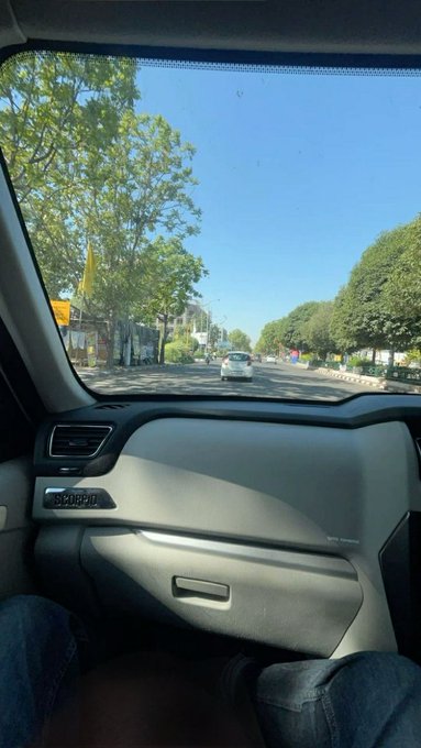 A view from inside a Jeep passenger seat looking out through the windshield at a tree-lined street with a white car driving ahead under a clear blue sky.