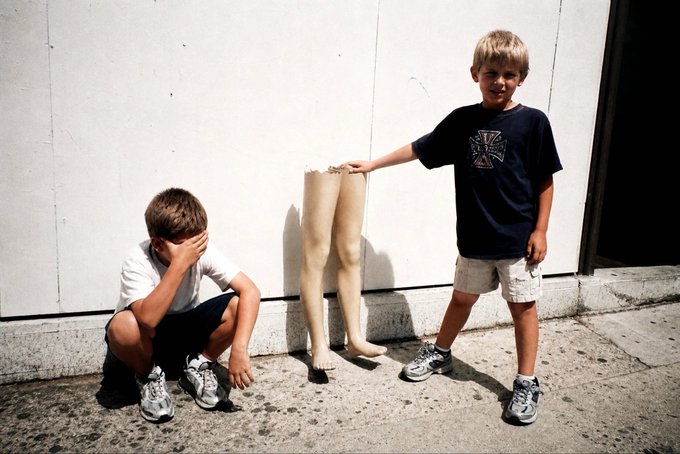 Two young boys posing on a sidewalk next to a pair of disembodied mannequin legs leaning against a white wall; one boy crouches with his hand over his face while the other stands smiling with his hand on the legs.