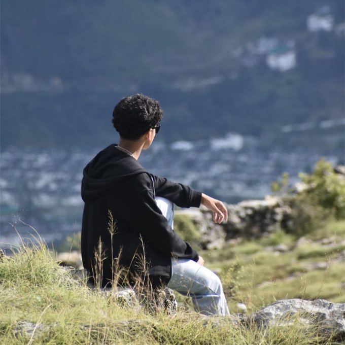 A person wearing a black hoodie and sunglasses sits on grassy terrain looking out over a vast, hazy mountainous landscape.